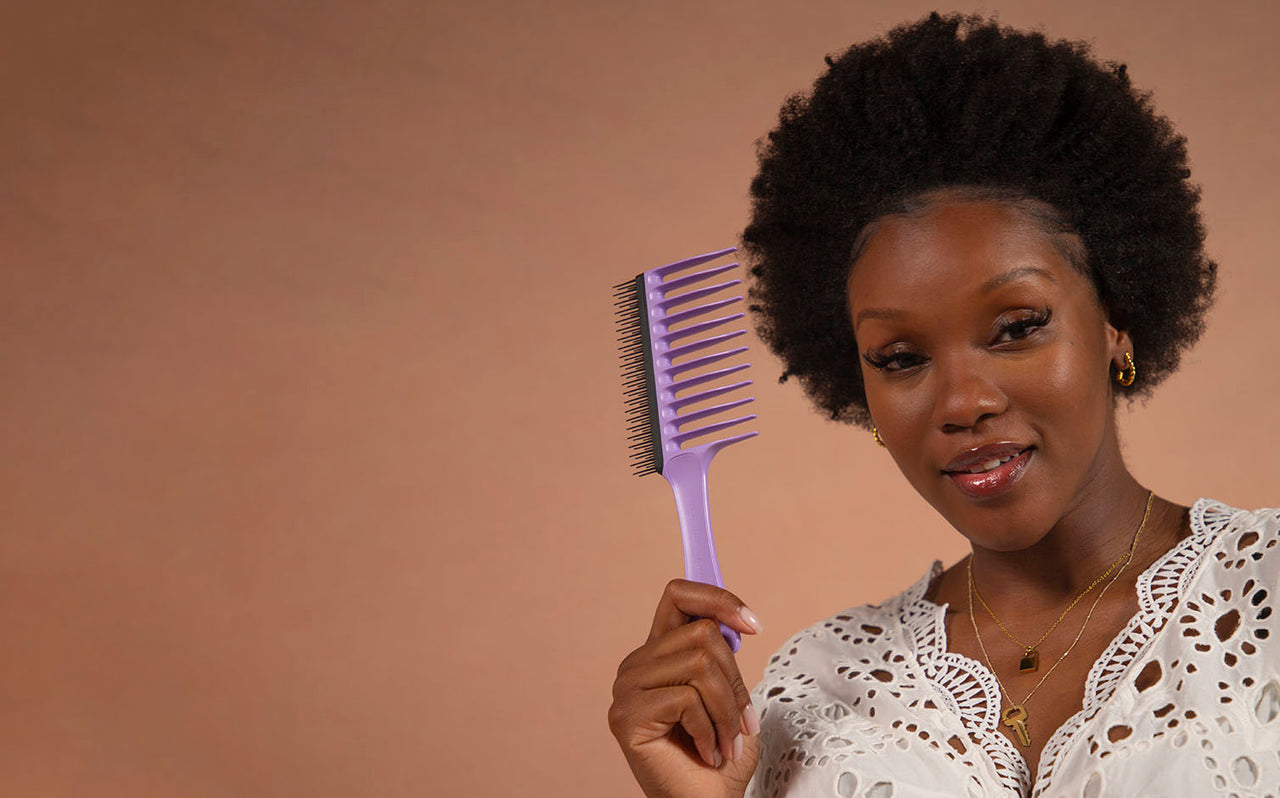 Smiling woman with natural afro hair holding a purple wide-tooth and pick comb against beige background