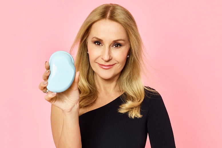 Smiling woman with blonde hair in black dress holding a light blue hairbrush against pink background
