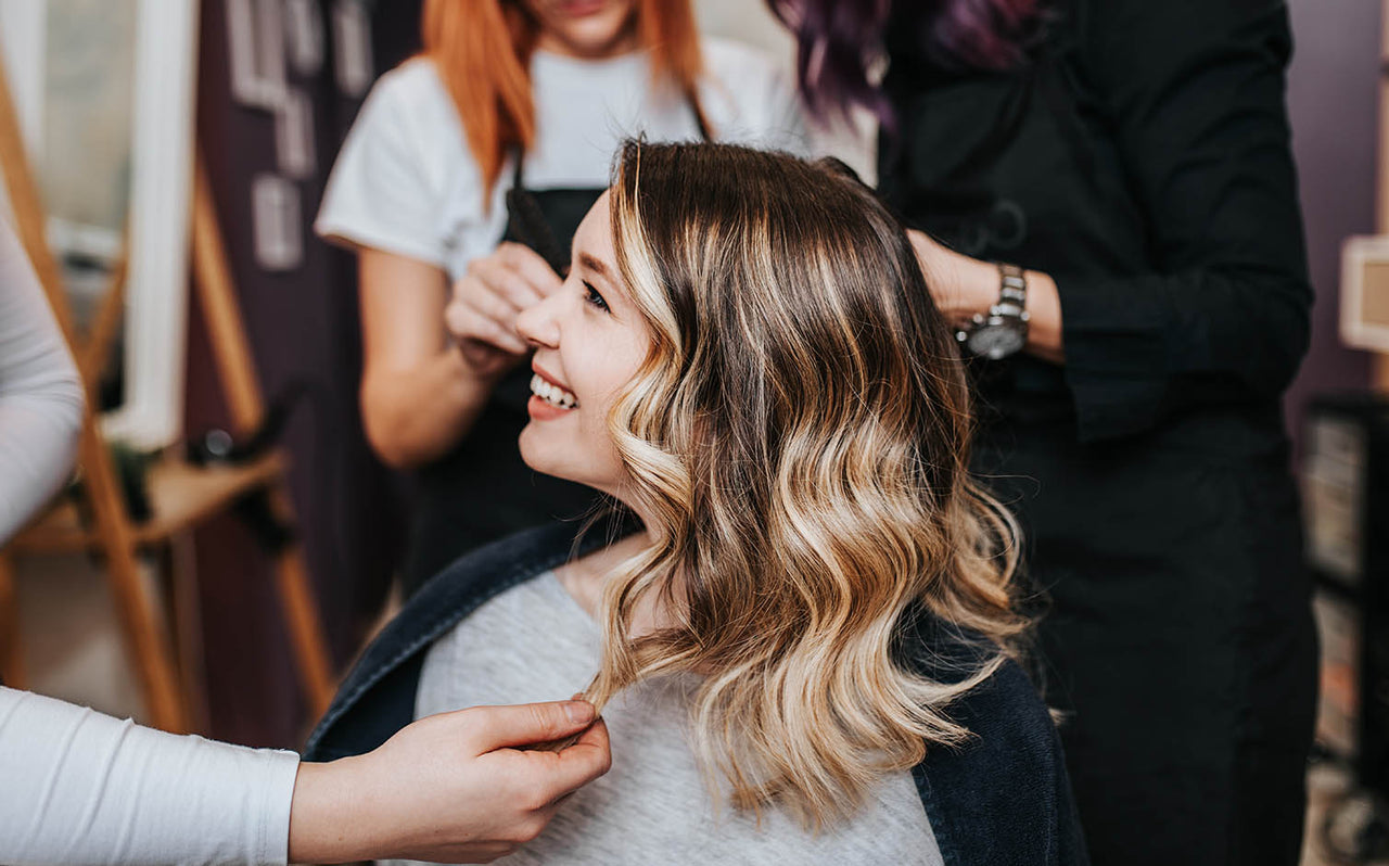 Smiling woman with highlighted wavy hair getting styled by hairdressers in salon