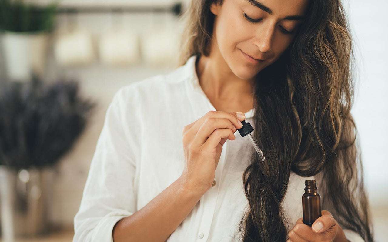 Woman in white shirt applying hair oil with dropper to long brown hair in bright room