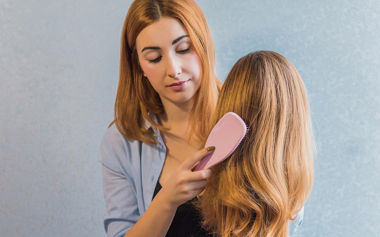 Woman brushing a long blonde wig with a pink hairbrush against a light blue background