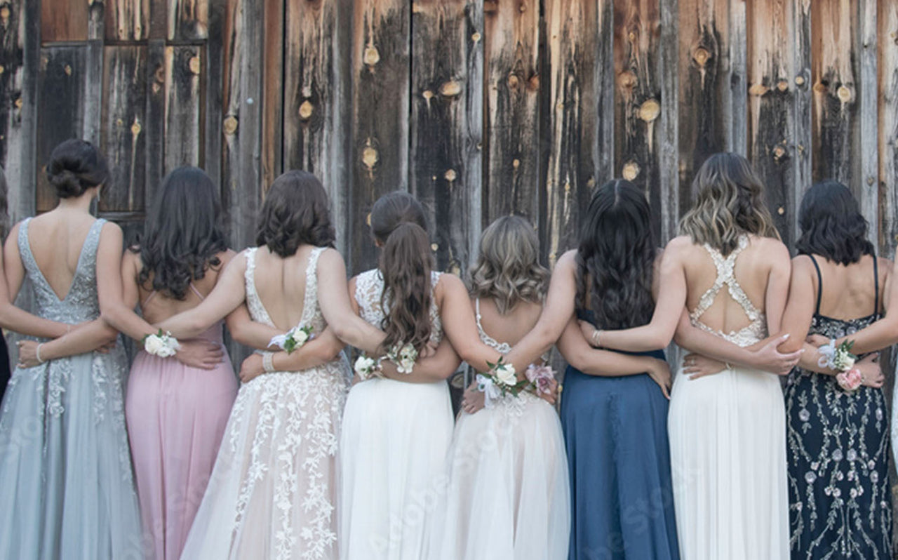 Eight women in elegant prom dresses with corsage bracelets, arms linked, standing against rustic wooden wall