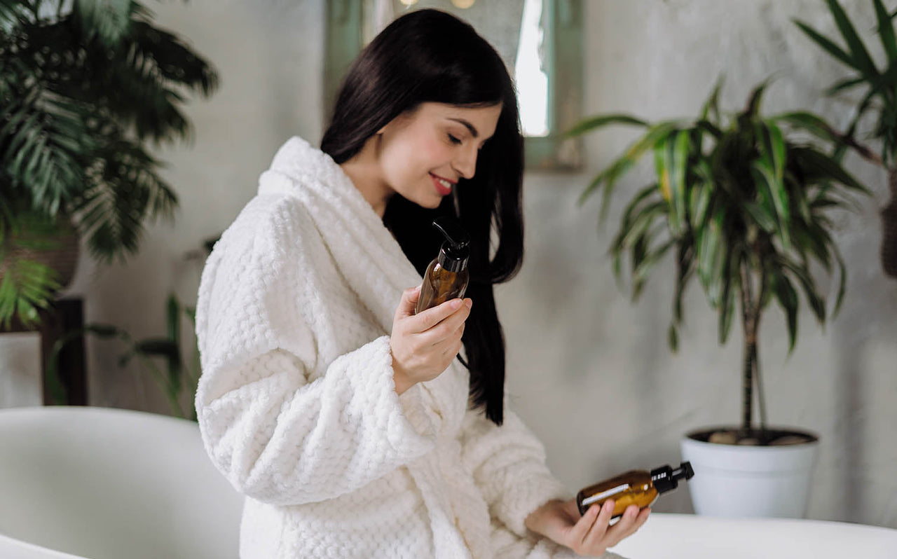 Woman in white bathrobe holding amber skincare bottles in a cozy bathroom with green plants