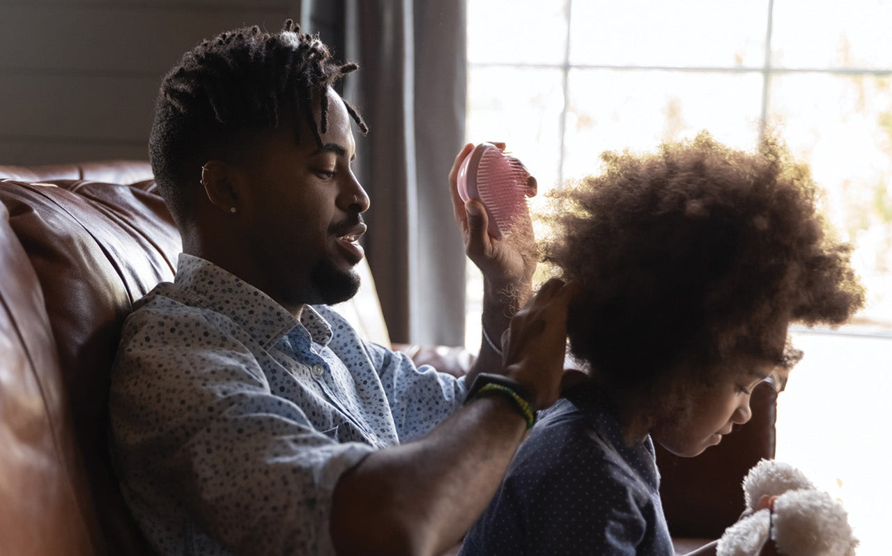 Man brushing young girl's curly hair while sitting on couch near window