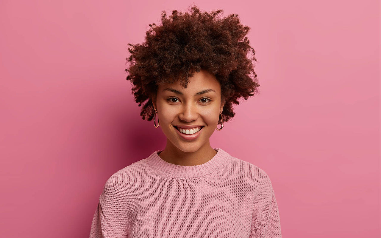 Smiling young woman with curly afro hair wearing pink sweater and hoop earrings against pink background