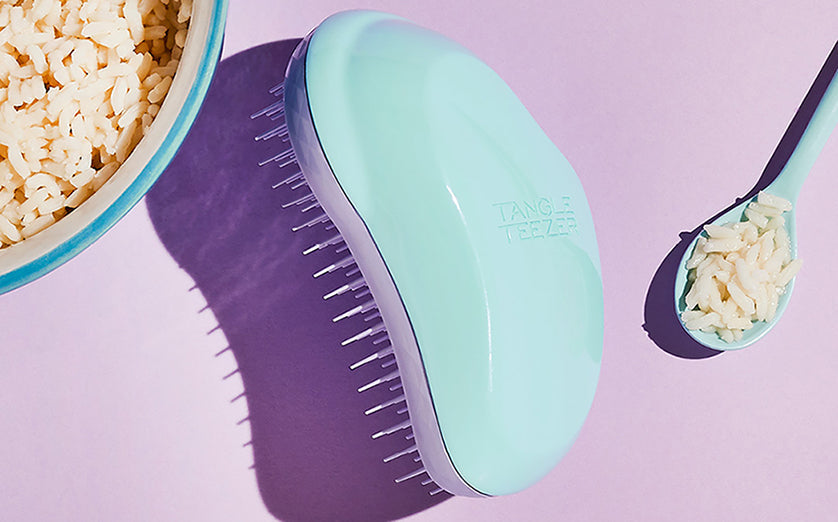 Mint green Tangle Teezer hairbrush next to a bowl and spoon of cooked rice on a pastel purple background