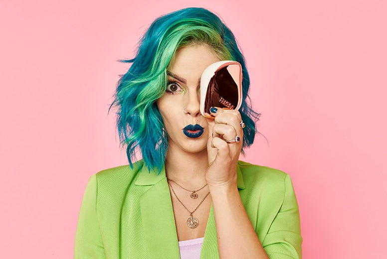 Young woman with blue-green hair, blue lipstick, and green blazer holding a pink facial cleansing device against pink background