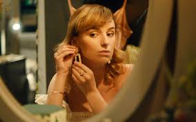 Young woman with blonde hair wearing earrings, looking in a round mirror in a softly lit room